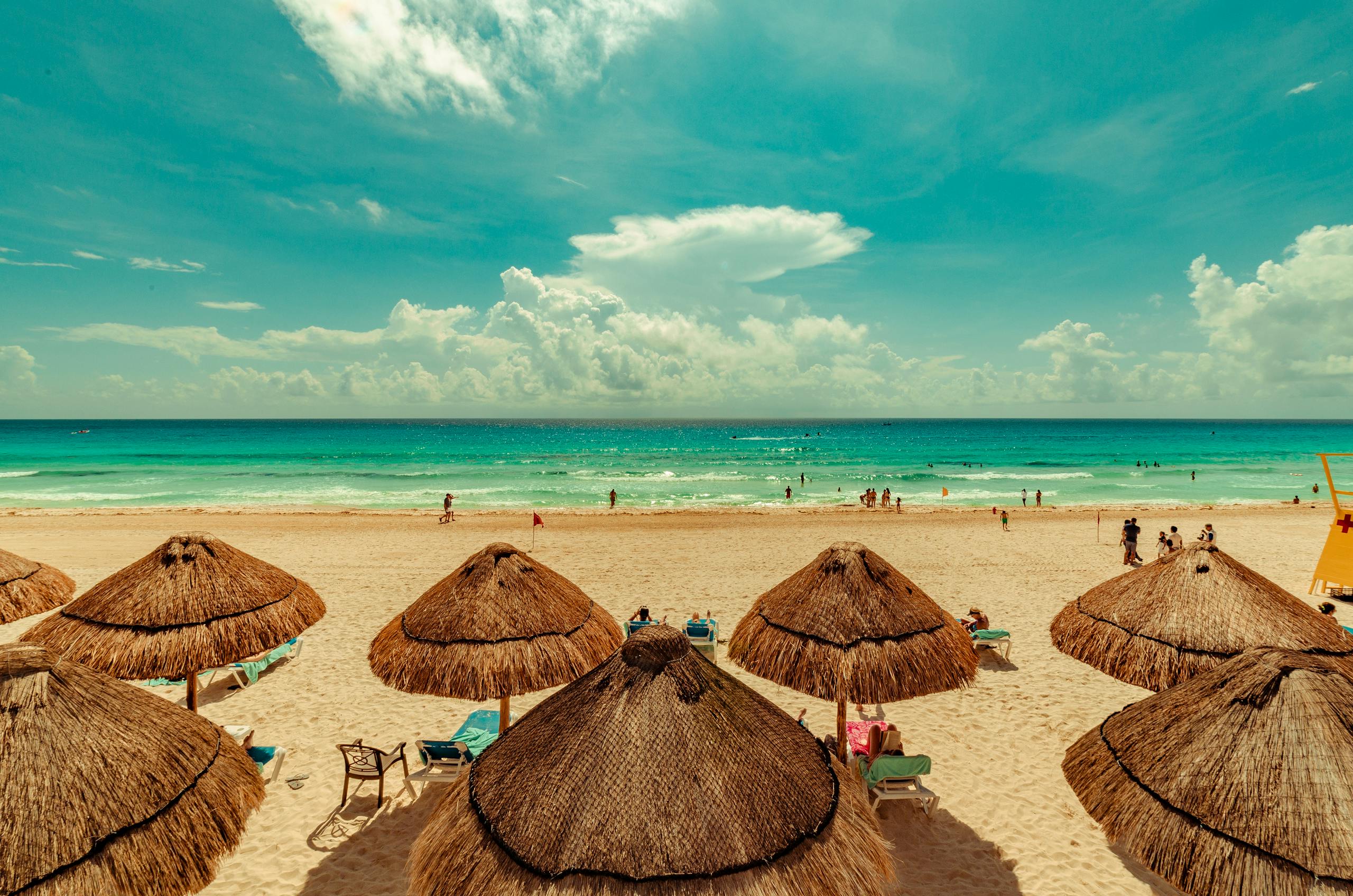 A vibrant beach scene in Cancun, Mexico with straw umbrellas and turquoise sea under a bright sky.